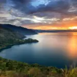 paket wisata danau toba: Pemandangan indah Danau Toba saat matahari terbenam dengan langit berwarna keemasan, dikelilingi perbukitan hijau yang menambah pesona wisata alam di Sumatera Utara.
