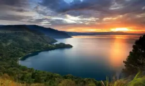 paket wisata danau toba: Pemandangan indah Danau Toba saat matahari terbenam dengan langit berwarna keemasan, dikelilingi perbukitan hijau yang menambah pesona wisata alam di Sumatera Utara.