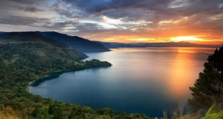 paket wisata danau toba: Pemandangan indah Danau Toba saat matahari terbenam dengan langit berwarna keemasan, dikelilingi perbukitan hijau yang menambah pesona wisata alam di Sumatera Utara.