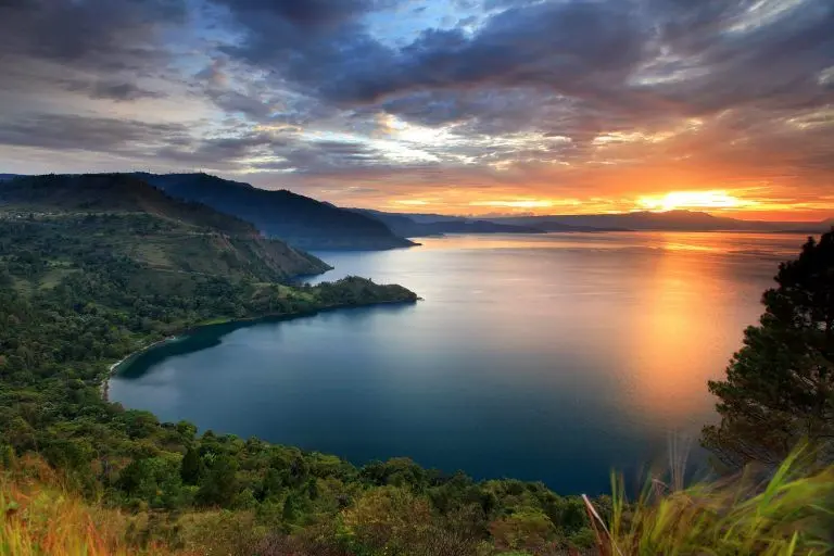 paket wisata danau toba: Pemandangan indah Danau Toba saat matahari terbenam dengan langit berwarna keemasan, dikelilingi perbukitan hijau yang menambah pesona wisata alam di Sumatera Utara.